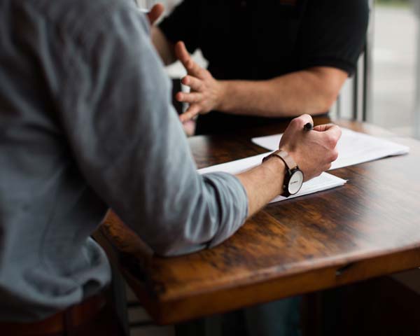 one man at desk wearing watch and holding pen above paper while man across from him gestures with his hands