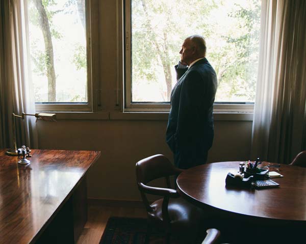 older man in suit standing in front of window inside of office on cell phone